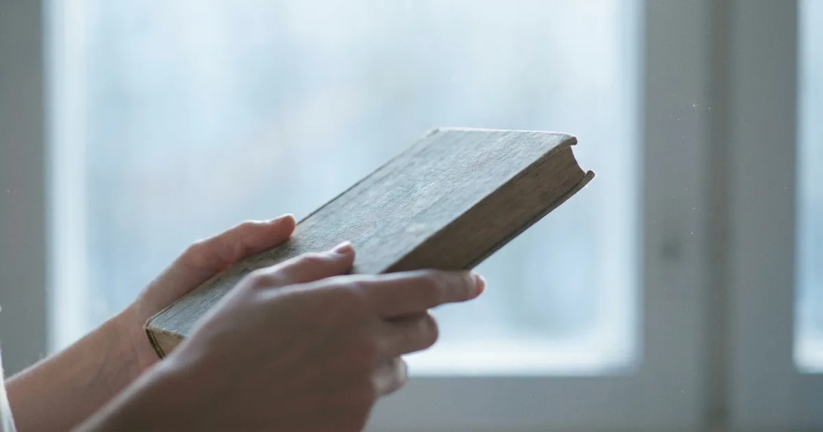 A person holding an old hardcover book near a window.
